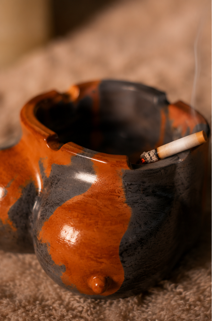 A ceramic novelty smoking ashtray with a glossy orange-brown and dark gray glaze, shown in a three-panel collage on a textured beige surface and wooden table. The product has a bulbous, organic shape with a small protruding nipple detail on one side and a lit cigarette resting inside the open top.