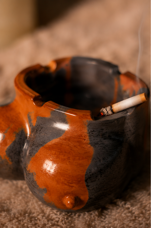 A ceramic novelty smoking ashtray with a glossy orange-brown and dark gray glaze, shown in a three-panel collage on a textured beige surface and wooden table. The product has a bulbous, organic shape with a small protruding nipple detail on one side and a lit cigarette resting inside the open top.