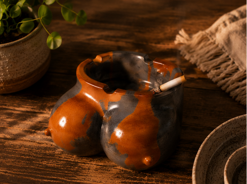 A ceramic novelty smoking ashtray with a glossy orange-brown and dark gray glaze, shown in a three-panel collage on a textured beige surface and wooden table. The product has a bulbous, organic shape with a small protruding nipple detail on one side and a lit cigarette resting inside the open top.