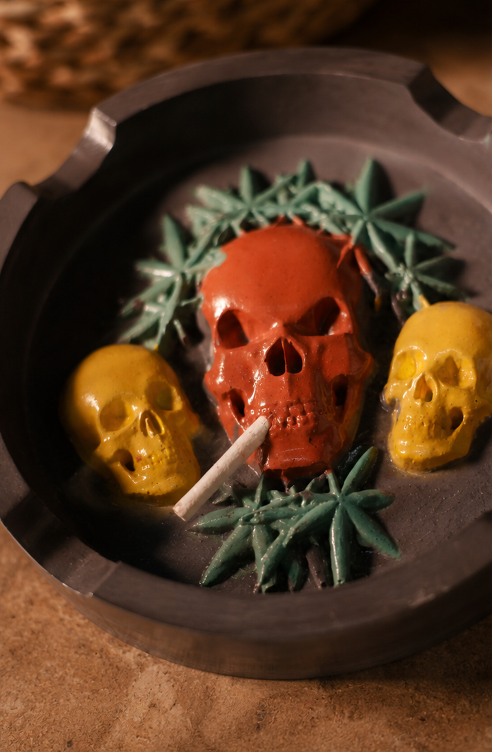 A triptych collage showing a decorative skull-shaped smoking ashtray in use. The left panel shows the ashtray empty, filled with a red skull, two yellow skulls, green leaf accents, and a white cigarette.