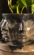 A ceramic planter shaped like a stylized human face, shown in a three-panel collage. The planter has a dark charcoal gray glaze with subtle greenish-blue undertones and holds a green leafy plant.