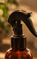 Collage showing an amber glass spray bottle with a black trigger sprayer in three warm-toned scenes: a close-up of the sprayer, the bottle standing on a wooden surface, and a hand spraying mist in a bedroom.